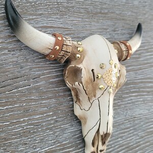 Peut inclure: Cr&acirc;ne de taureau en c&eacute;ramique blanche avec des accents dor&eacute;s et des lani&egrave;res en cuir marron. Le cr&acirc;ne est d&eacute;cor&eacute; de strass et d'un motif en m&eacute;tal dor&eacute;.