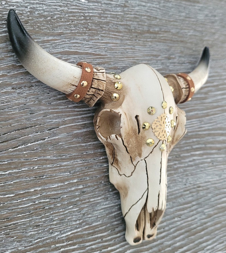 Peut inclure: Cr&acirc;ne de taureau en c&eacute;ramique blanche avec des accents dor&eacute;s et des lani&egrave;res en cuir marron. Le cr&acirc;ne est d&eacute;cor&eacute; de strass et d'un motif en m&eacute;tal dor&eacute;.