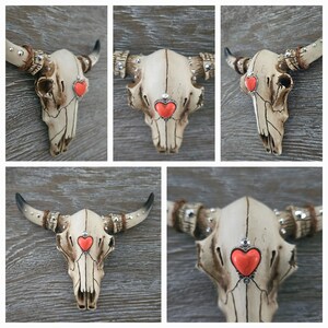 May include: A white ceramic bull skull with rhinestones and a red heart on the forehead. The skull is mounted on a wooden wall.