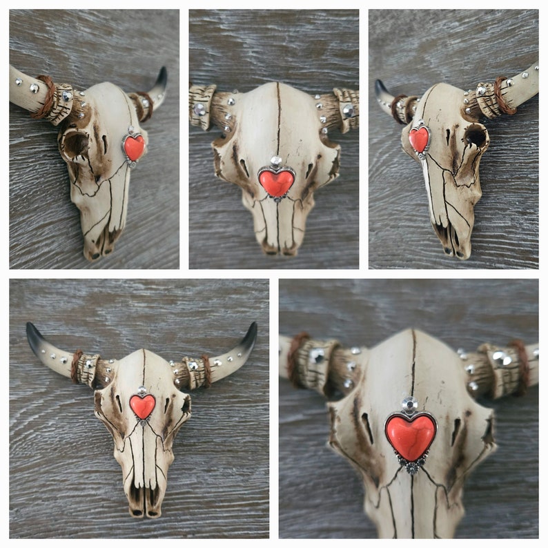 May include: A white ceramic bull skull with rhinestones and a red heart on the forehead. The skull is mounted on a wooden wall.