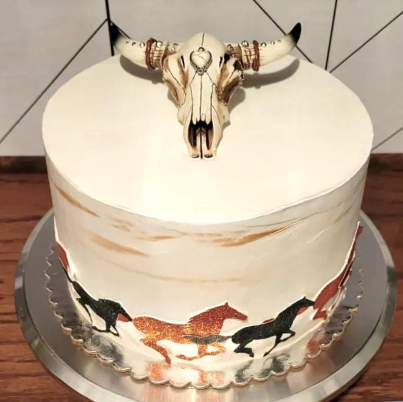 May include: A white cake decorated with a brown and white bull skull and a border of brown and black horses running around the bottom of the cake.