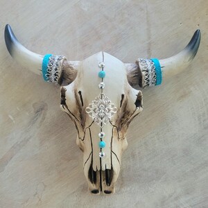 May include: A white ceramic bull skull with turquoise and silver accents. The skull is decorated with turquoise beads, silver filigree, and turquoise and silver bands around the horns.