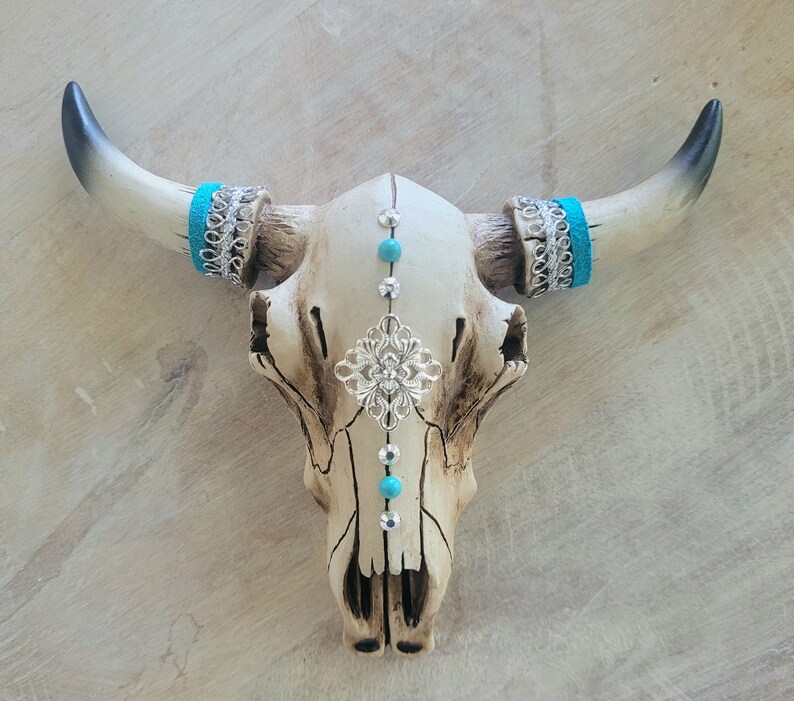 May include: A white ceramic bull skull with turquoise and silver accents. The skull is decorated with turquoise beads, silver filigree, and turquoise and silver bands around the horns.