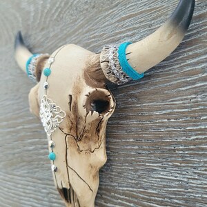 May include: A white ceramic bull skull with turquoise and silver accents. The skull is decorated with a silver filigree design and turquoise beads. The horns are black and white.