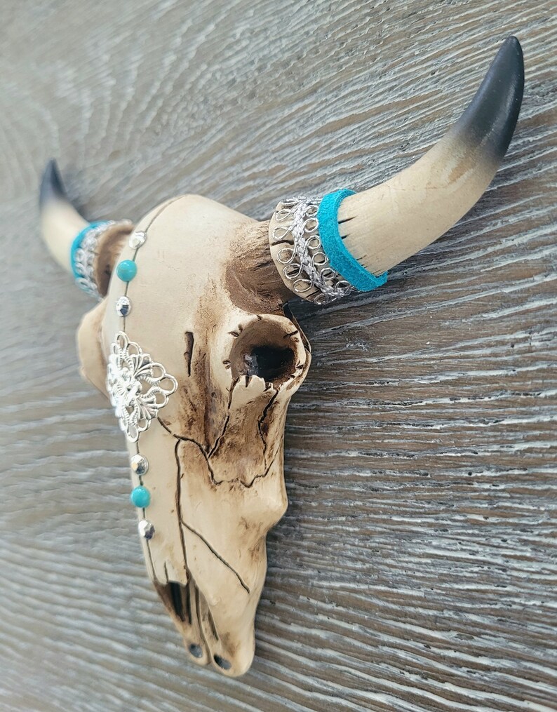 May include: A white ceramic bull skull with turquoise and silver accents. The skull is decorated with a silver filigree design and turquoise beads. The horns are black and white.