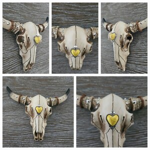 May include: A white ceramic bull skull with gold accents and a yellow heart-shaped stone in the center of the forehead. The skull is mounted on a wooden wall.