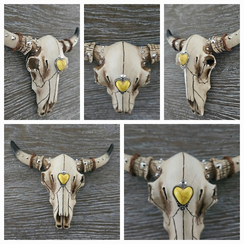 May include: A white ceramic bull skull with gold accents and a yellow heart-shaped stone in the center of the forehead. The skull is mounted on a wooden wall.