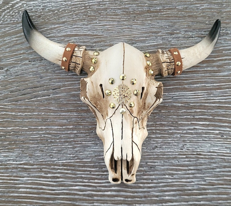 Peut inclure: Un cr&acirc;ne de taureau en c&eacute;ramique blanche avec des accents dor&eacute;s et des lani&egrave;res en cuir marron. Le cr&acirc;ne est d&eacute;cor&eacute; d'un motif en &eacute;toile dor&eacute;e.