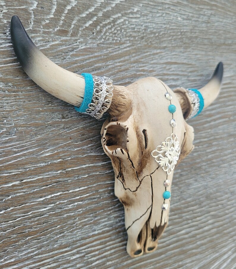 May include: White ceramic bull skull with turquoise and silver accents. The skull has a distressed finish and is decorated with turquoise beads and silver filigree.