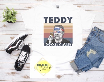 May include: White t-shirt featuring a graphic of Theodore Roosevelt with an American flag bandana and sunglasses, holding a drink. The text "TEDDY BOOZEDEVELT" is printed above and below the image. The shirt is displayed with jeans and sneakers.