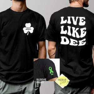May include: A black t-shirt featuring a white shamrock graphic on the front chest with the text "DEE'S ANGELS" inside. The back displays the white text "LIVE LIKE DEE" in a bold font. A sleeve detail shows a green awareness ribbon with "BK".