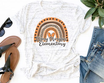 May include: A white and gray marbled t-shirt featuring a rainbow design with orange and leopard print accents. The text "Mary Morgan Elementary" is printed below the rainbow in black. The shirt is displayed with sunglasses, sandals, and jeans.
