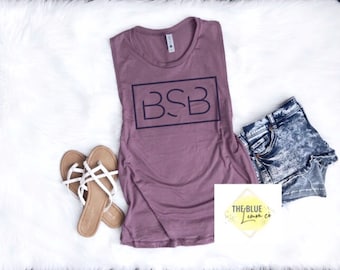 May include: A mauve-colored tank top with the letters "BSB" inside a black rectangle, paired with denim shorts and sandals. The sandals are brown with white straps. The items are arranged on a white, fluffy surface.