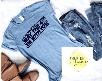 May include: Light blue t-shirt with the text "STAR WARS DAY MAY THE 4TH BE WITH YOU" in a black rectangular design. The shirt is paired with distressed blue jeans and brown boots.