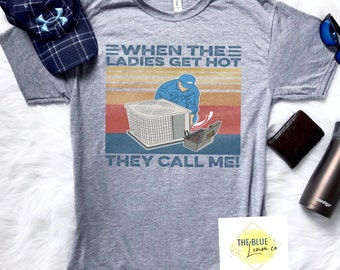 May include: A heather gray t-shirt with the text "WHEN THE LADIES GET HOT THEY CALL ME!" and a graphic of an HVAC technician working on an air conditioning unit. A blue and navy plaid baseball cap is visible.