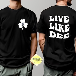 May include: Black t-shirt with white text and graphics. The front features a white shamrock with the words "LIVE DEE" and a pink ribbon. The back of the shirt displays the words "LIVE LIKE DEE" in a wavy font.