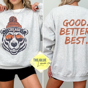 May include: A light gray sweatshirt featuring a bear wearing sunglasses and a beanie with "Chicago" on it. The back of the sweatshirt has the words "GOOD. BETTER. BEST." in orange. The sweatshirt is a casual style.