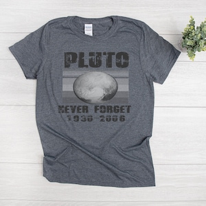 May include: A dark gray t-shirt featuring a graphic of Pluto with the text "PLUTO" above and "NEVER FORGET 1930-2006" below. The graphic includes an image of Pluto and horizontal gray stripes.