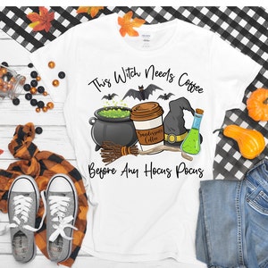 May include: White t-shirt with a Halloween-themed graphic. The design includes a witch's cauldron, coffee cup, witch hat, and the text "This Witch Needs Coffee Before Any Hocus Pocus". The shirt is paired with jeans and sneakers.