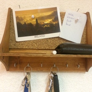 May include: A wooden key holder with a corkboard and a shelf. The corkboard has a postcard with a picture of a town in Ireland and a handwritten note that says "Milch, Wasser, Brot". There are two sets of keys hanging from the hooks.