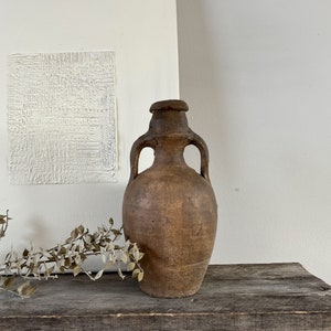 May include: A rustic brown clay jug with two handles, sitting on a wooden surface. The jug has a slightly rounded body and a wide mouth.