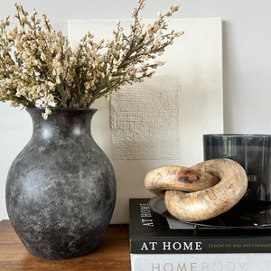 May include: A black vase with a textured surface holds a bouquet of dried white flowers. The vase is sitting on a wooden table next to a stack of books. The top book is titled "At Home" and the bottom book is titled "Homebody". A wooden knot sculpture sits on top of the books.
