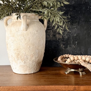 Peut inclure: Un grand vase blanc cassé texturé avec deux poignées contient des branches vertes et feuillues. Un petit bol en laiton antique avec des perles de bois et un pompon est posé sur une surface en bois. Le fond est un mur sombre et texturé.