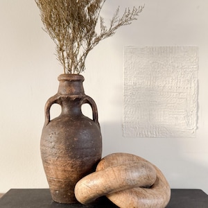 May include: A weathered brown ceramic vase with two handles contains dried, light brown branches. A wooden decorative chain is in front of the vase. A white textured wall art piece is in the background.