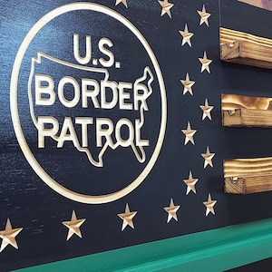 May include: Wooden wall art featuring a black background with gold stars and a green stripe. The center displays a circle with the text "U.S. Border Patrol" and a map of the United States. Includes three wooden shelves.