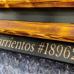 May include: A wooden sign with a black background and a gold inscription that reads "Barrientes #18962". The sign has a rustic, distressed look and is made of multiple layers of wood.