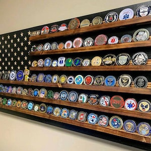 May include: A wall-mounted wooden display rack featuring horizontal shelves for organizing circular challenge coins. The back panel is painted black with a pattern of light-colored stars, resembling a flag design, holding numerous metallic coins.