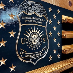 May include: A wooden American flag with a navy blue field and gold stars. The flag features a Homeland Security Investigations emblem and the words "Special Agent." The wooden stripes are a natural wood color.