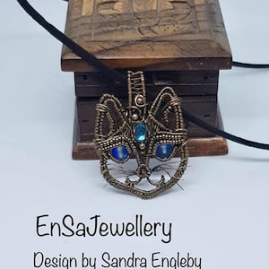 May include: A wire-wrapped cat pendant with blue gemstone eyes. The pendant is on a wooden box with the text "EnSaJewellery Design by Sandra Engleby".