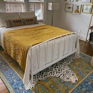 May include: A bedroom with a white metal bed frame, a mustard yellow quilt, and a patterned rug. The rug features a blue border and a leopard print design. The room has natural light from the window.