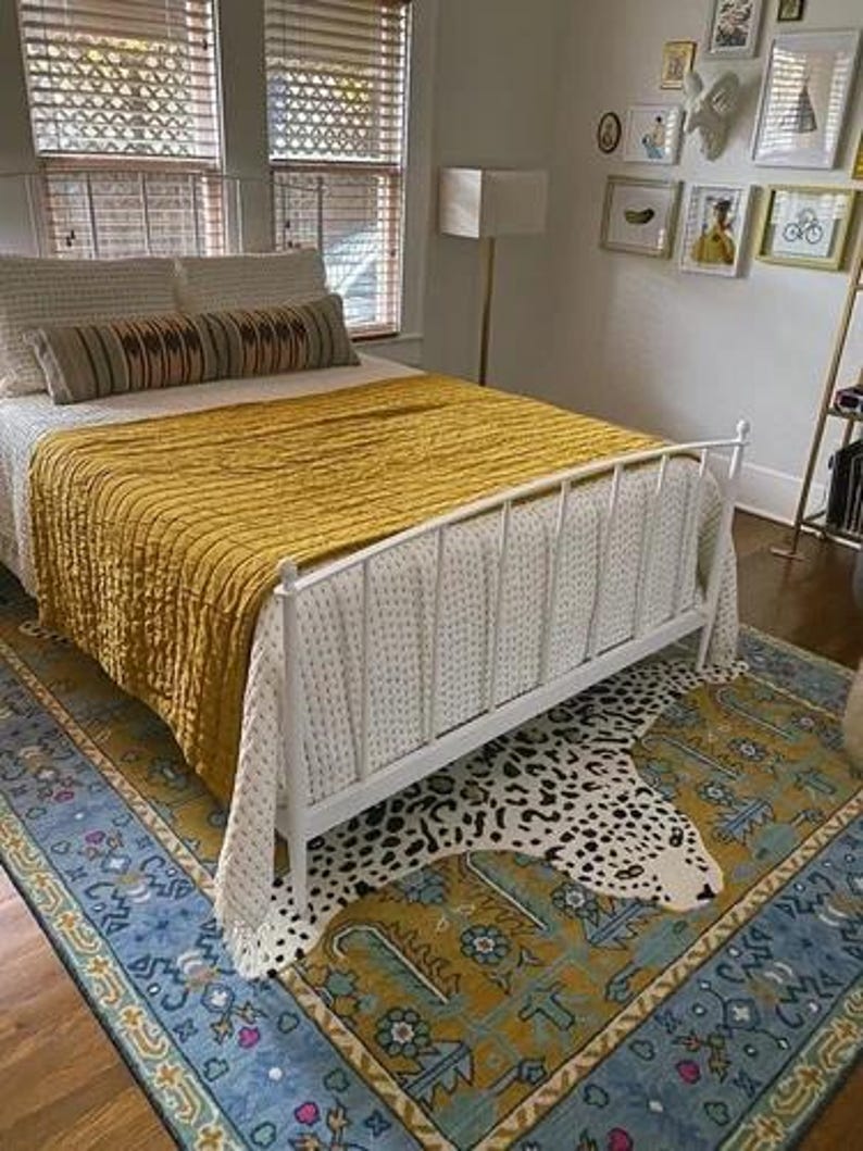 May include: A bedroom with a white metal bed frame, a mustard yellow quilt, and a patterned rug. The rug features a blue border and a leopard print design. The room has natural light from the window.