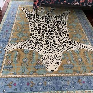 May include: A large area rug with a leopard print design in the center, surrounded by a patterned border in shades of blue, gold, and beige. The rug is placed on a dark wooden floor.