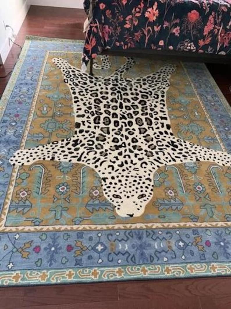 May include: A large area rug with a leopard print design in the center, surrounded by a patterned border in shades of blue, gold, and beige. The rug is placed on a dark wooden floor.