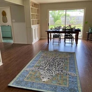 May include: A rectangular area rug with a leopard print design in the center. The rug has a blue border with a floral pattern and is placed on a hardwood floor. The room has a dining table and chairs.
