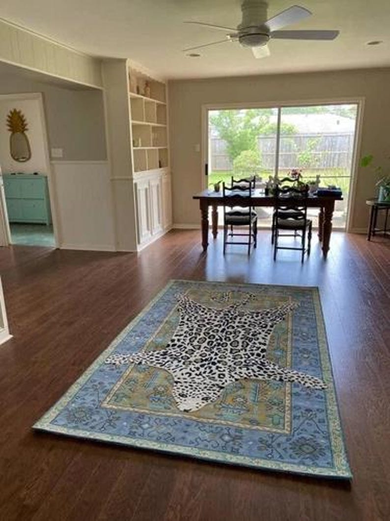 May include: A rectangular area rug with a leopard print design in the center. The rug has a blue border with a floral pattern and is placed on a hardwood floor. The room has a dining table and chairs.