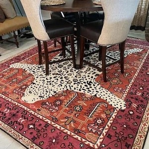 May include: A decorative rug with a leopard print design in the center, surrounded by a red and orange floral border. The rug is placed under a dark wood table and chairs with cream-colored backs and dark brown seats.