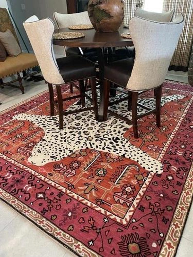 May include: A decorative rug with a leopard print design in the center, surrounded by a red and orange floral border. The rug is placed under a dark wood table and chairs with cream-colored backs and dark brown seats.