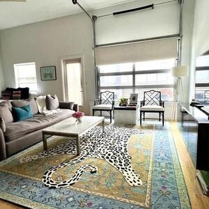 May include: A modern living room with a large brown sofa, a patterned rug with a leopard design, and a coffee table. The room has large windows, two black chairs, and a flat-screen TV.