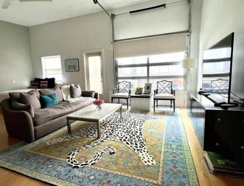 May include: A modern living room with a large brown sofa, a patterned rug with a leopard design, and a coffee table. The room has large windows, two black chairs, and a flat-screen TV.