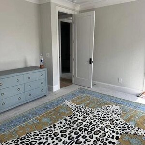 May include: A bedroom featuring a large area rug with a leopard print design. The rug is primarily tan and blue with a white and black leopard pattern. A light blue dresser is on the left, and a white door is open in the background.