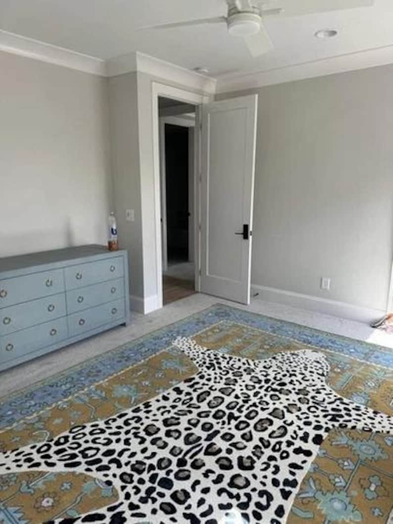 May include: A bedroom featuring a large area rug with a leopard print design. The rug is primarily tan and blue with a white and black leopard pattern. A light blue dresser is on the left, and a white door is open in the background.