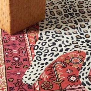May include: A woven leather ottoman in a warm brown hue sits next to a rug with a leopard print design. The rug also features a red and beige patterned border and a red central field with floral motifs.