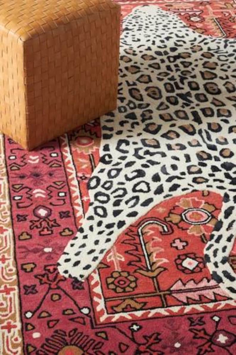 May include: A woven leather ottoman in a warm brown hue sits next to a rug with a leopard print design. The rug also features a red and beige patterned border and a red central field with floral motifs.