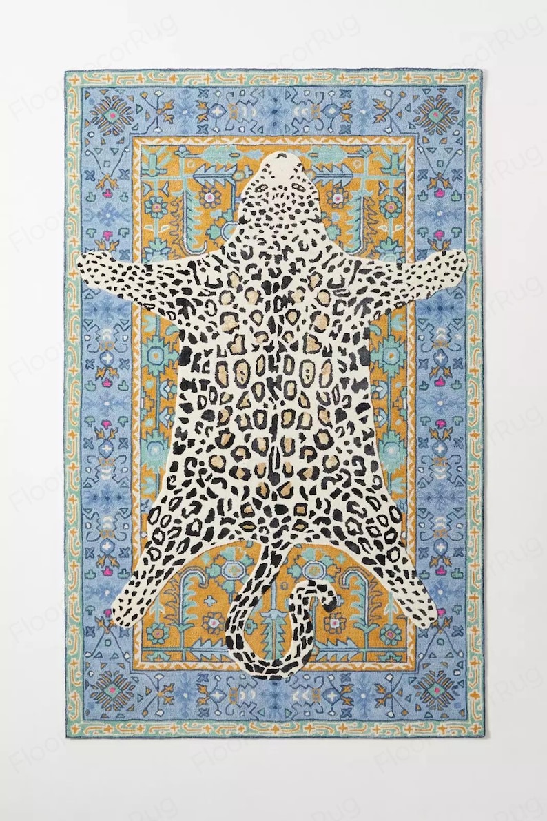 May include: A rectangular rug with a leopard skin design in black and tan spots. The rug is set against a blue and gold patterned border with floral and geometric motifs. The rug is a decorative floor covering.