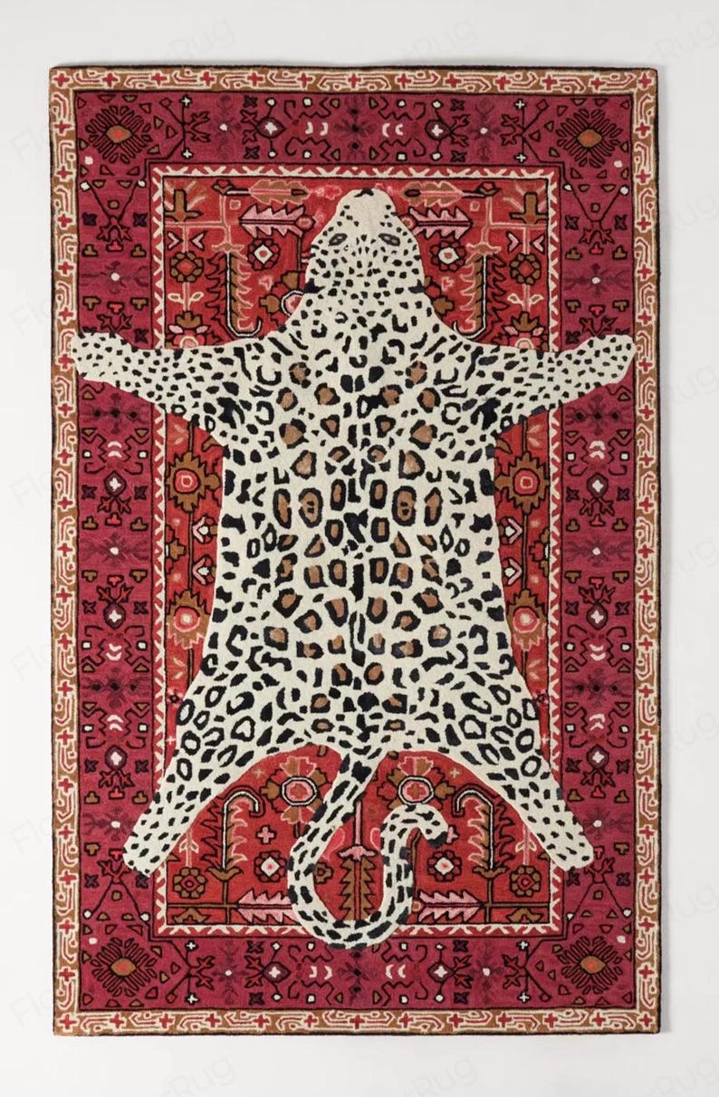 May include: A rectangular rug with a leopard skin design in the center, surrounded by a red and brown patterned border. The leopard print features black and brown spots on a cream background. The rug has a decorative, tribal-inspired design.