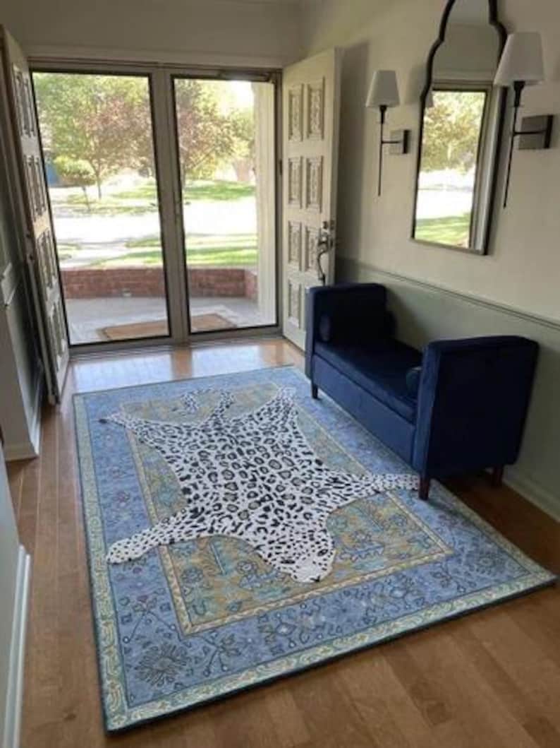 May include: A blue area rug with a detailed border and a leopard print rug on top. The leopard print rug is white with black spots. The room has a blue bench, a mirror, and a door.